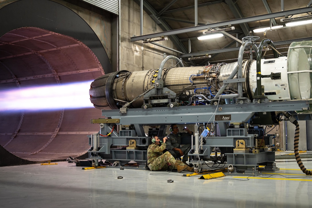 Two airmen shine flashlights at the underside of a jet engine blasting afterburner into a tunnel at an indoor facility.