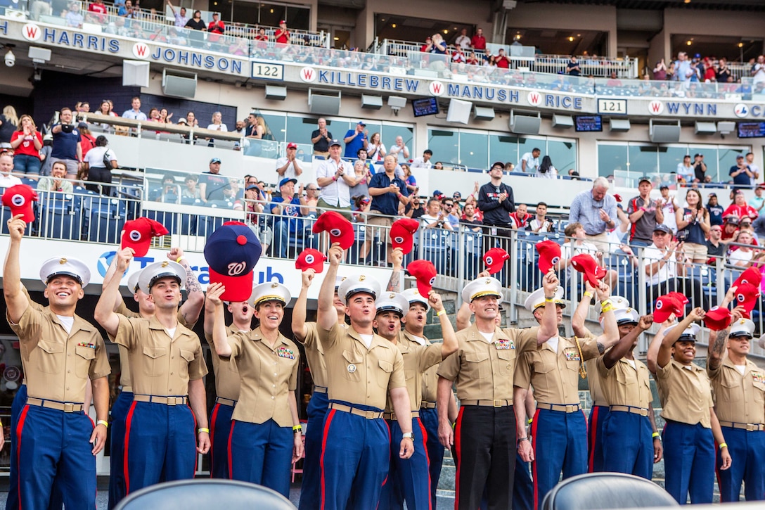 Marines in tan shirts and blue pants with a red stripe stand in a stadium holding team ballcaps in the air; fans in stands behind them applaud.