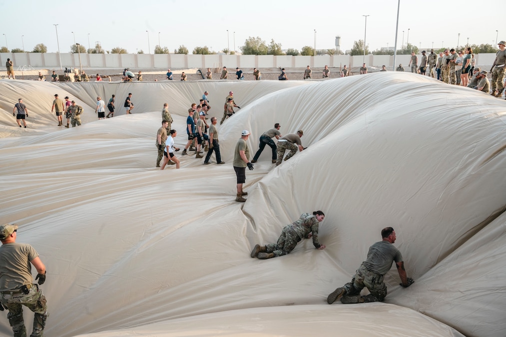 Airmen flatten a large beige liner by standing, crawling and walking on it outdoors.