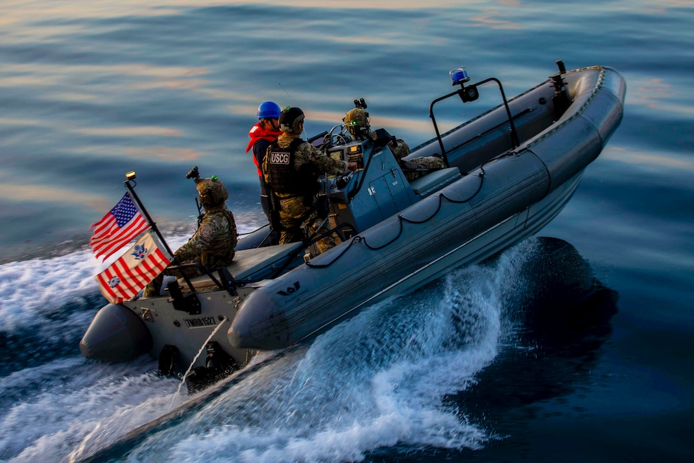Coast Guardsmen and sailors are seen riding in a small boat with two flags creating a wake in a smooth body of water.
