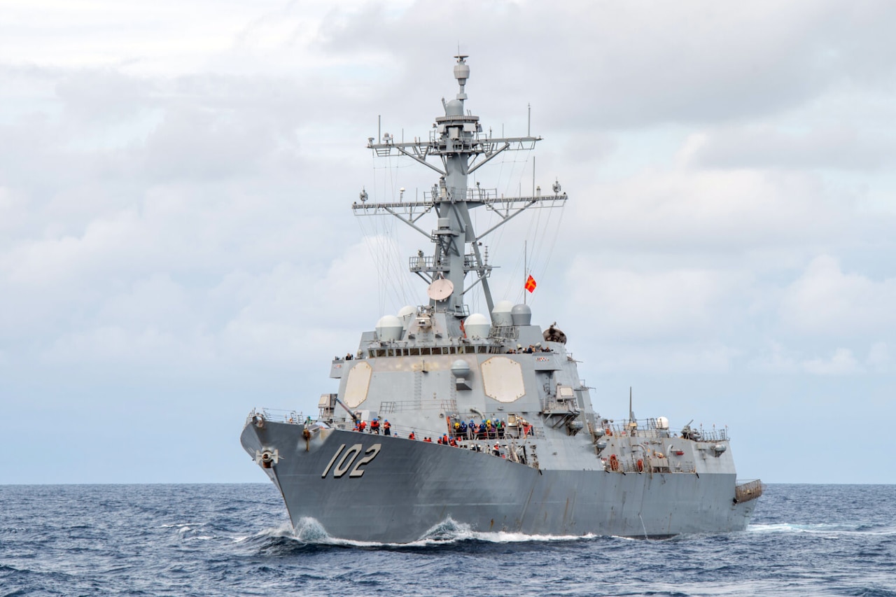 A large naval vessel moves through the ocean. The number 102 is painted on its side.