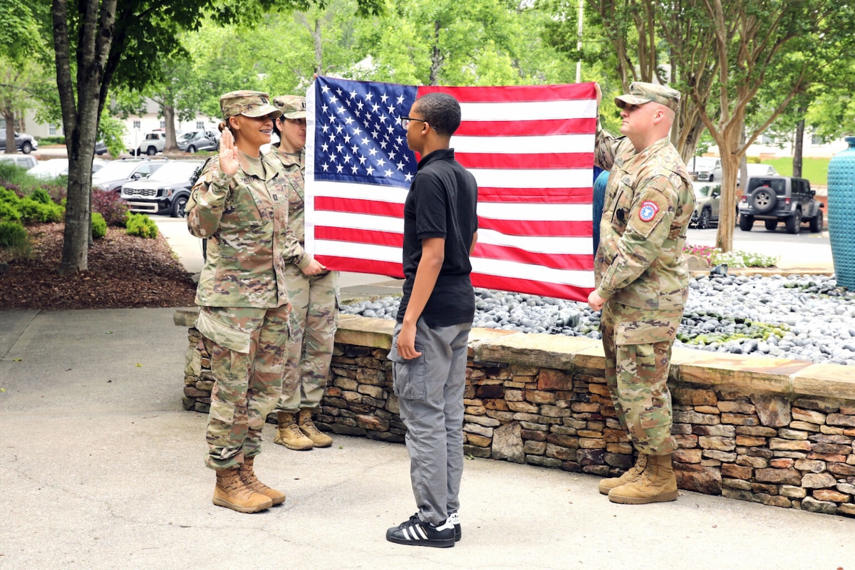 Outdoors, a woman in a military uniform and a young man in civilian clothing face each other and hold up their right hands. Behind them, two uniformed soldiers hold up a U.S. flag.