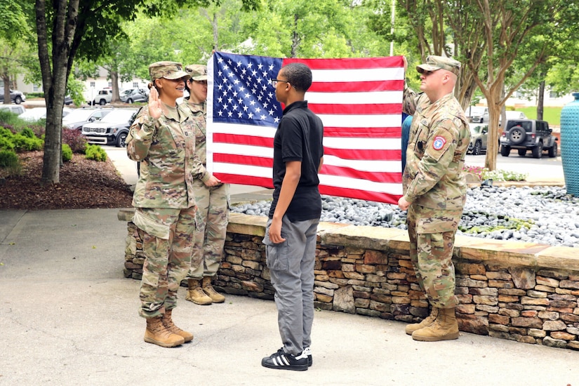 Outdoors, a woman in a military uniform and a young man in civilian clothing face each other and hold up their right hands. Behind them, two uniformed soldiers hold up a U.S. flag.