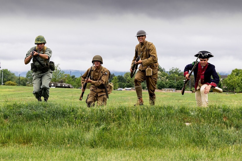 Four soldiers in historical uniforms run up a grassy hill while holding rifles.