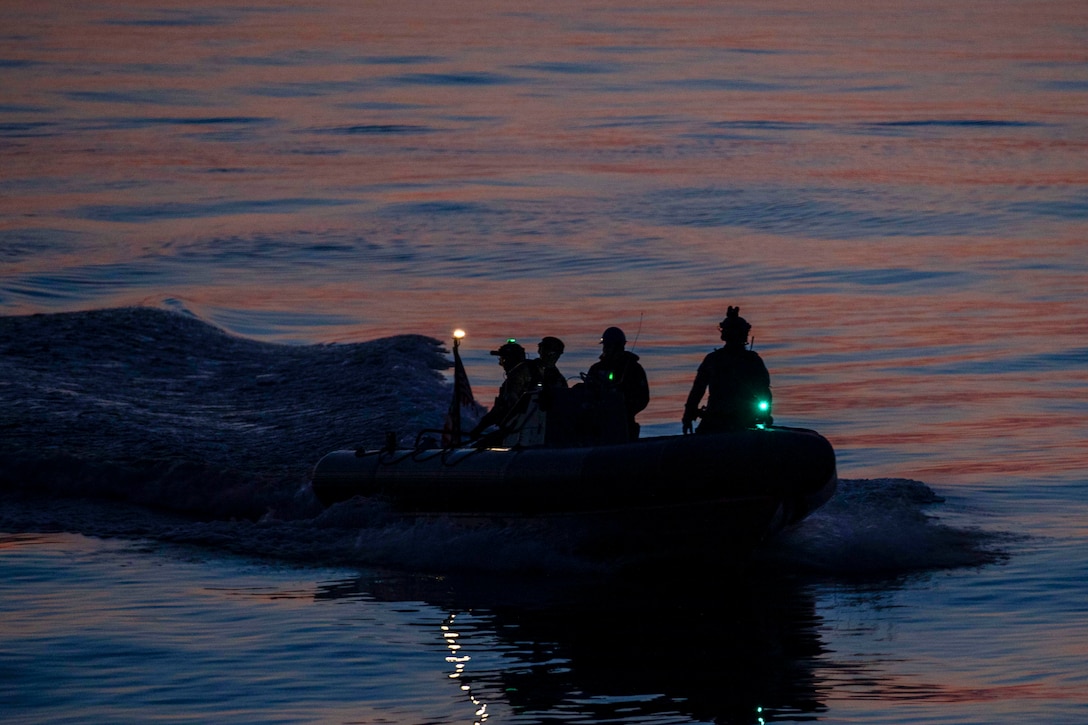 Coast Guardsmen and sailors are seen in silhouette riding in a small boat with lights creating a wake in a smooth body of water.