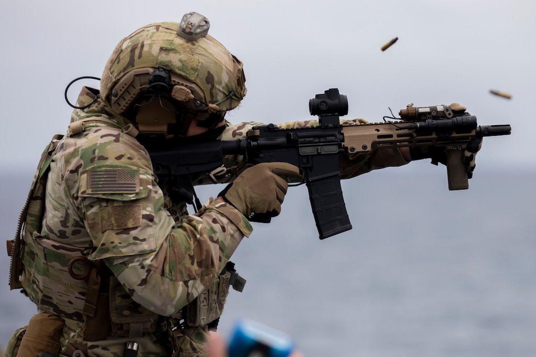 A Coast Guardsmen in combat gear fires a rifle with the ocean in the background.