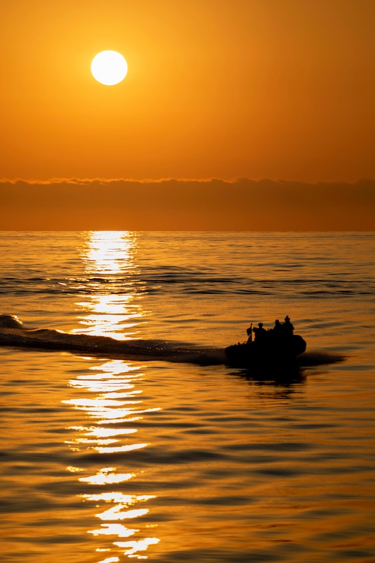 Coast Guardsmen and sailors are seen in silhouette riding in a small boat creating a wake in a smooth body of water with the sun and an orange sky in the background.