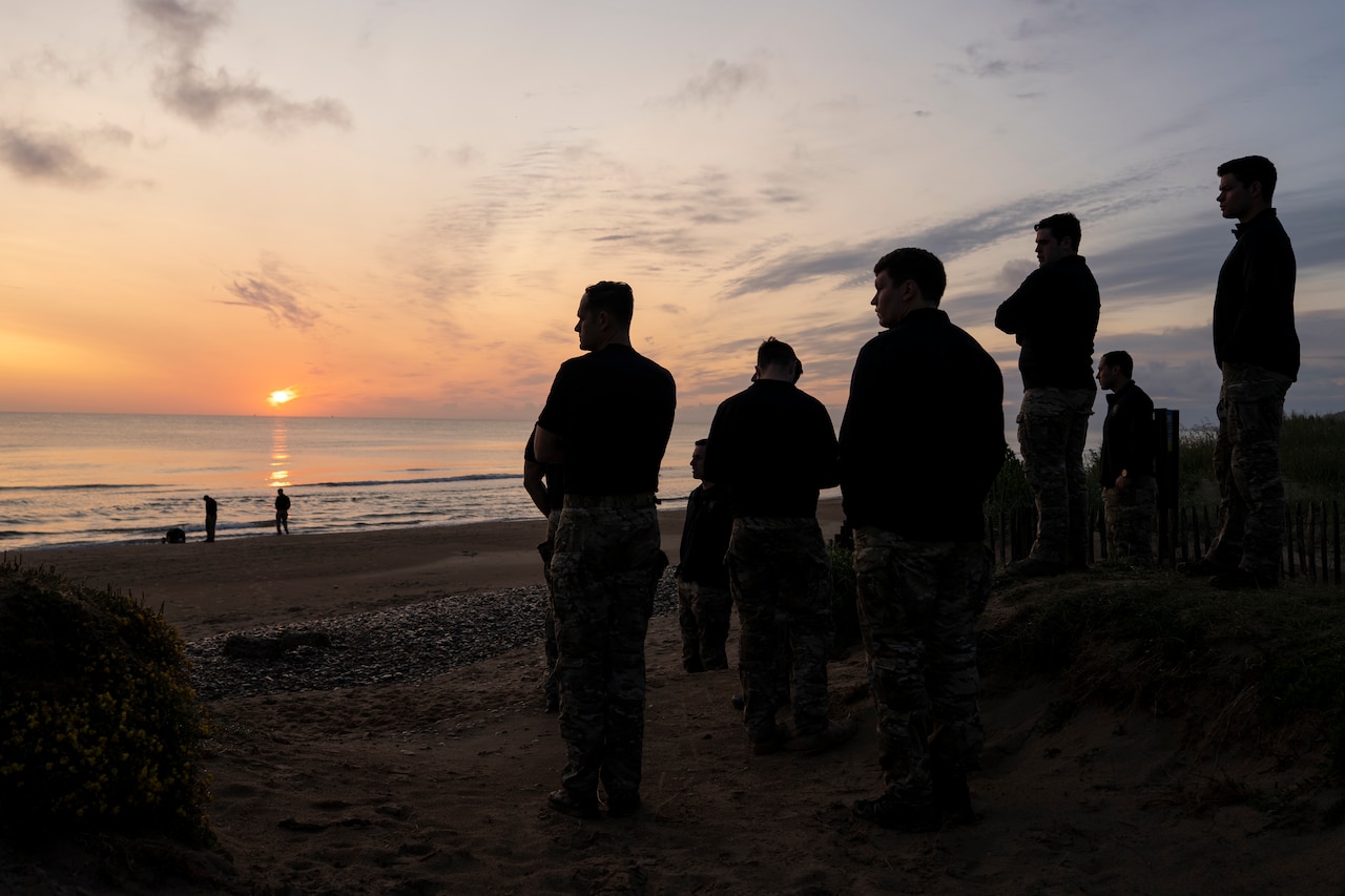Troops stand on a beach as the sun rises in the distance.