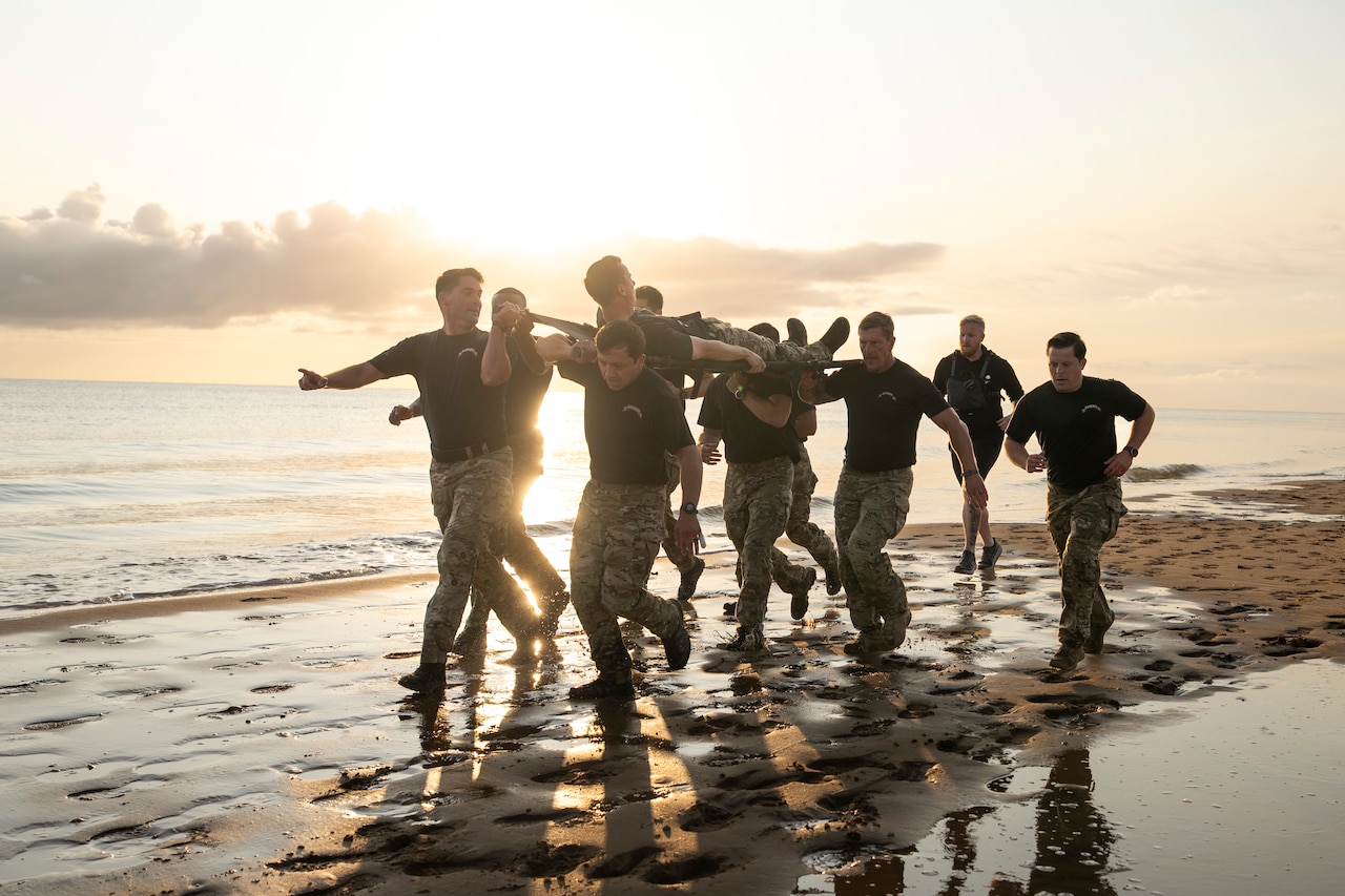 Soldiers in camouflage long pants and T-shirts run on a beach and carry a person lying on a stretcher.