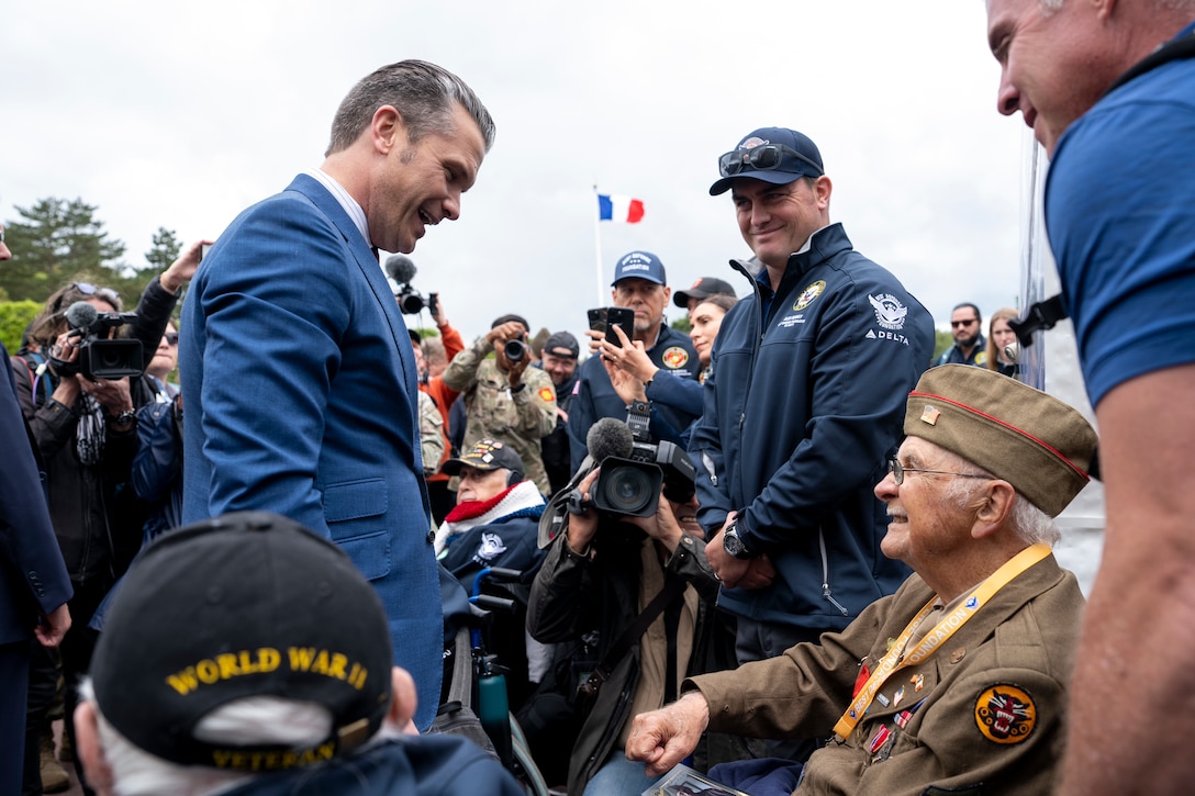 A man in a suit standing interacts with a veteran sitting as a crowd surrounding them looks on.