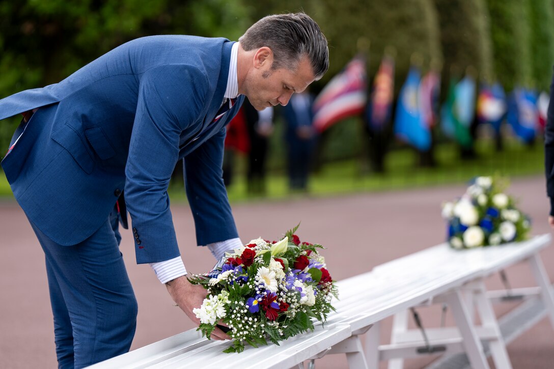 A person in a suit lays a wreath with flowers on a bench outdoors with another wreath and flags in the background.