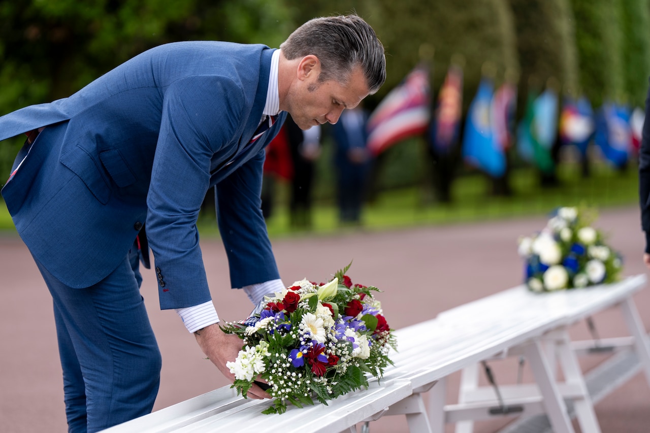 A person in a suit lays a wreath with flowers on a bench outdoors with another wreath and flags in the background.