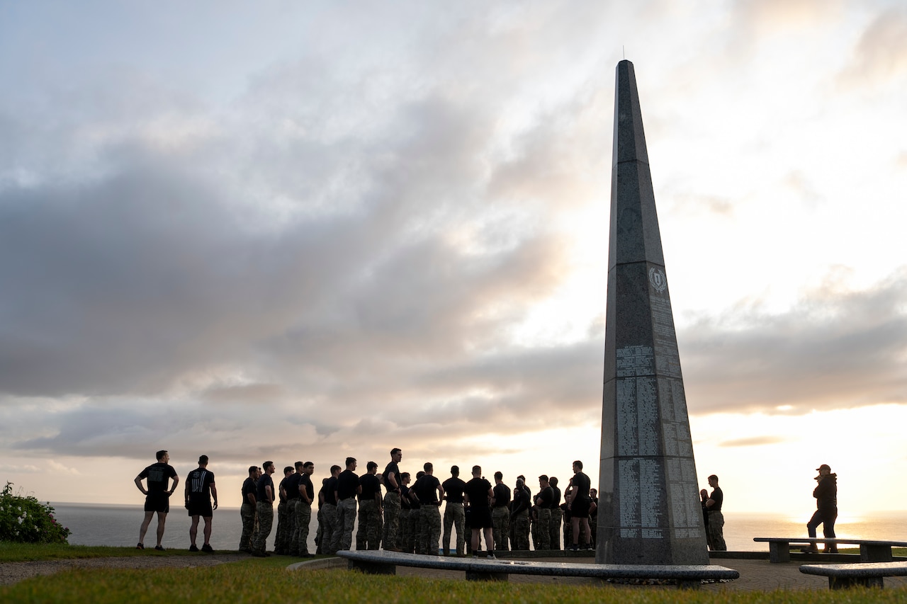 Troops stand near a monument on a cloudy day outdoors with the sea in the background.