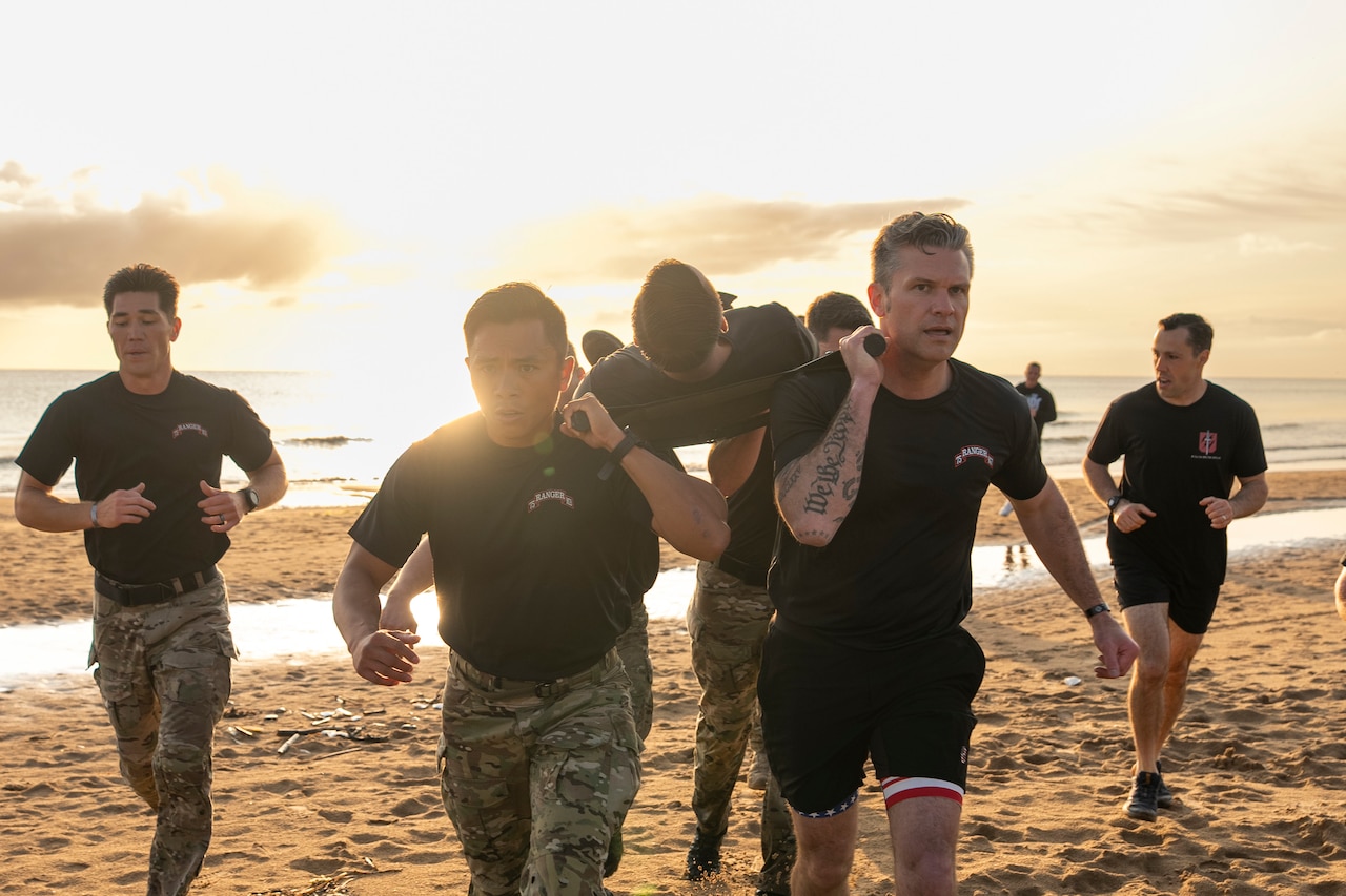 Soldiers in camouflage long pants and T-shirts run on a beach with another person in shorts and a T-shirt and carry a person lying on a stretcher.