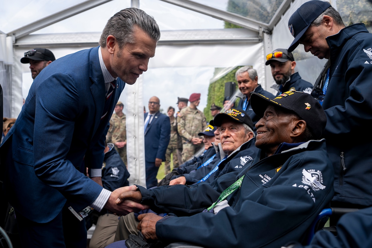 A man wearing a suit leans over to shake hands with a veteran sitting down. Other veterans sit in a line.