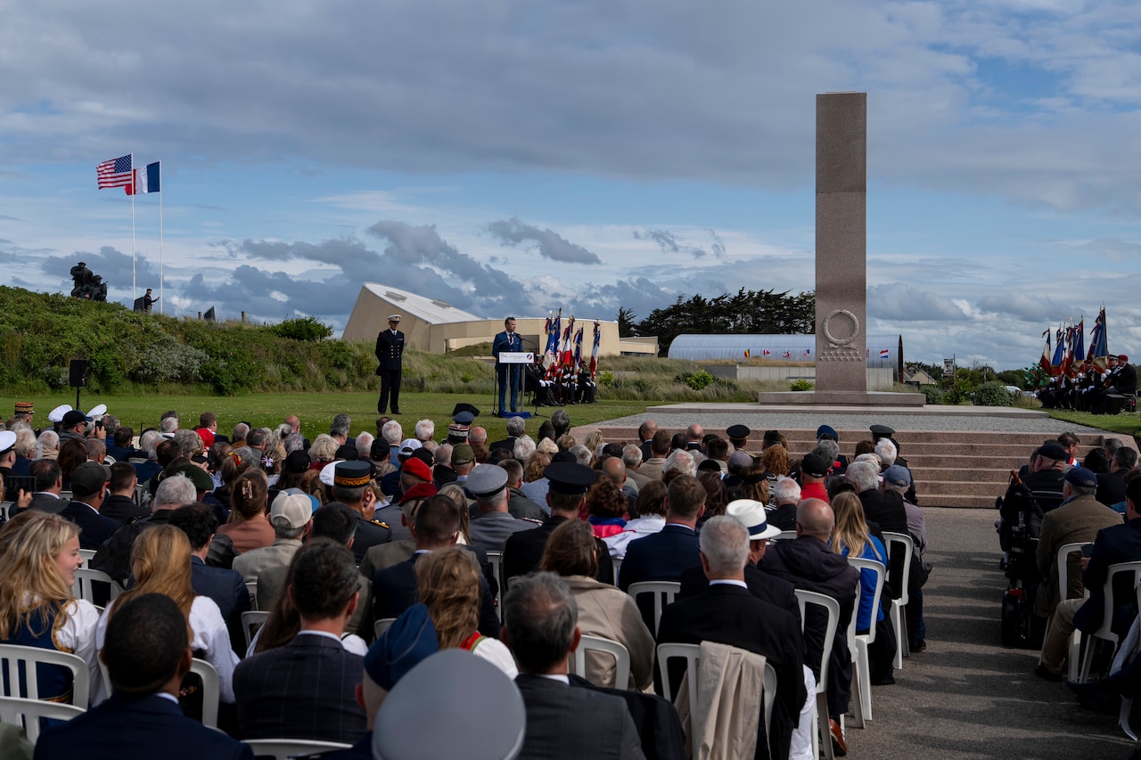 A person at a lectern to the left of a monument speaks to a seated audience outdoors with French and American flags in the background.