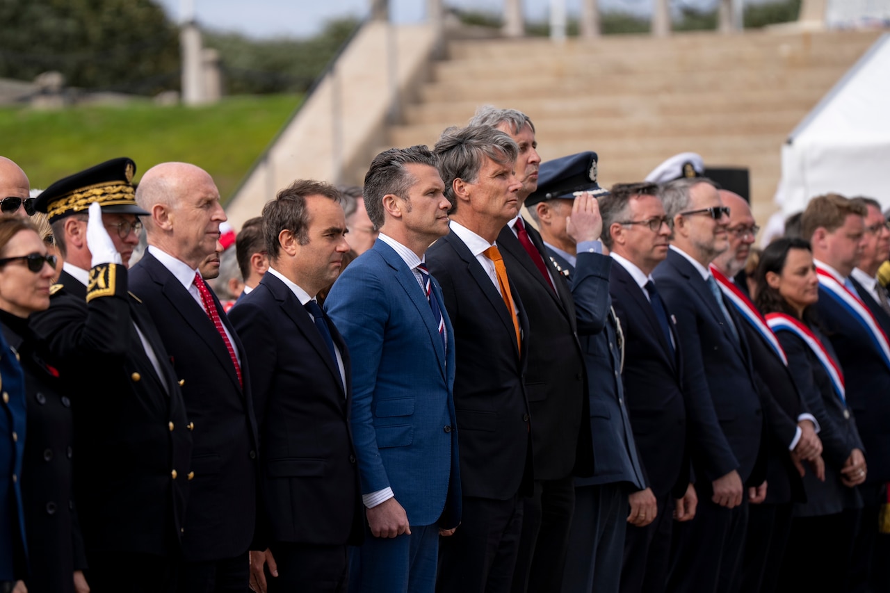 Standing people line up in a row outdoors with a staircase and grassy hill in the background.