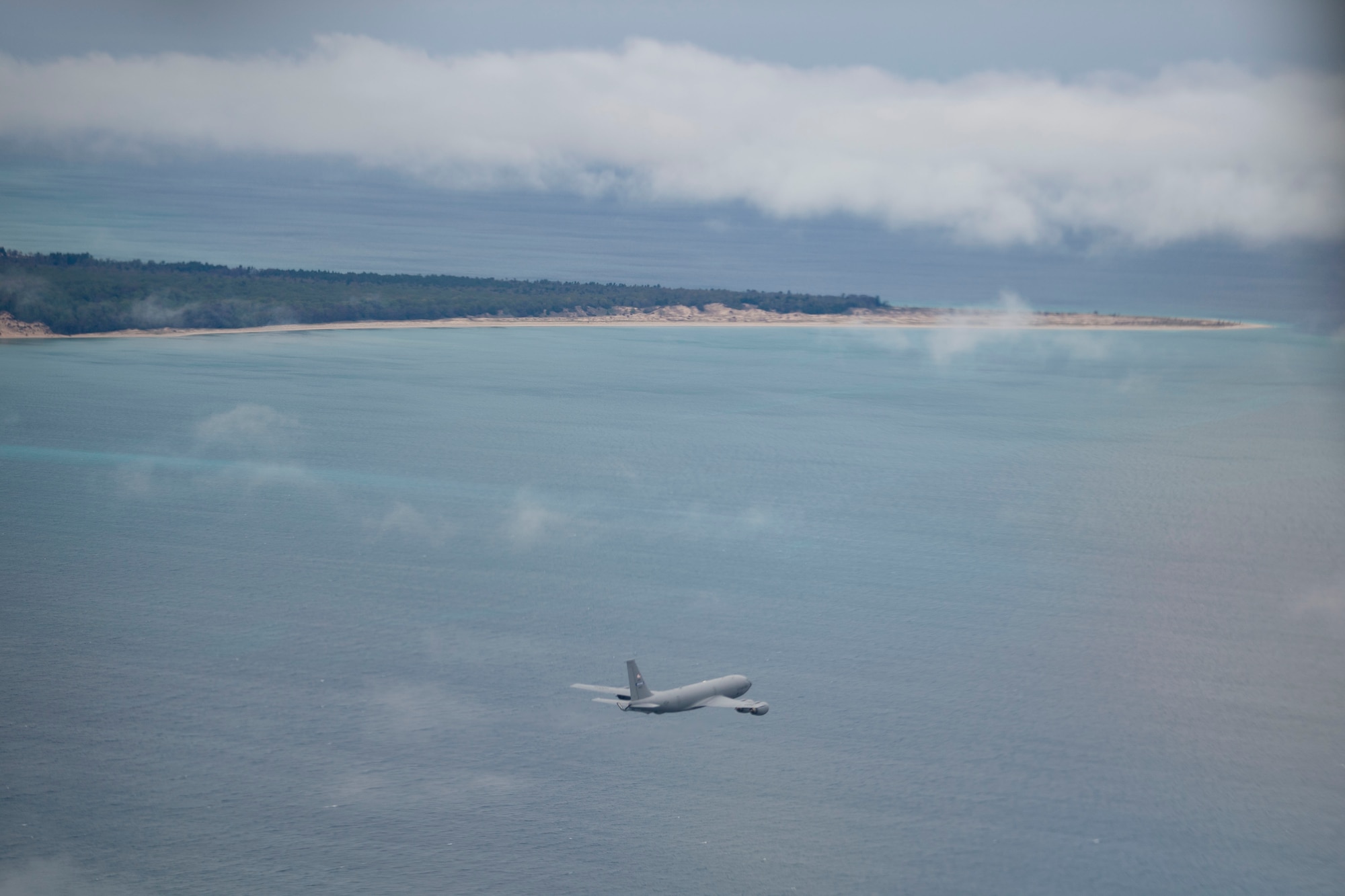 A KC-135R Stratotanker flies over water with beach and trees visible in the distance
