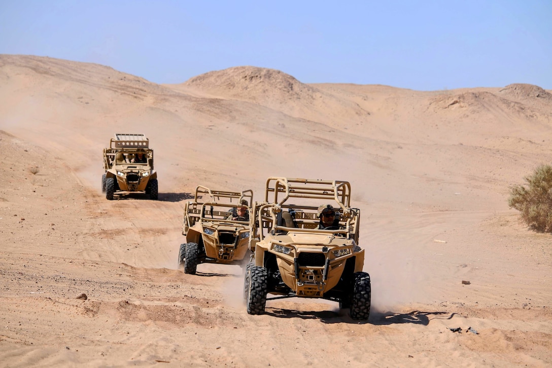 Three tan, open vehicles drive in line down a dry desert embankment.