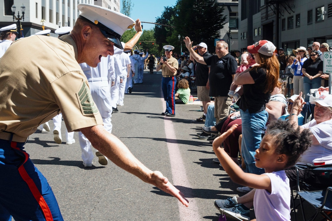 A Marine reaches out for a high-five from a child spectator during a parade with dozens of people in the background.