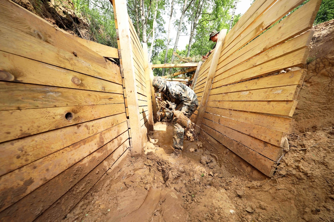 A service member shovels mud in a trench as a fellow service member watches from above with a wooded area in the background.