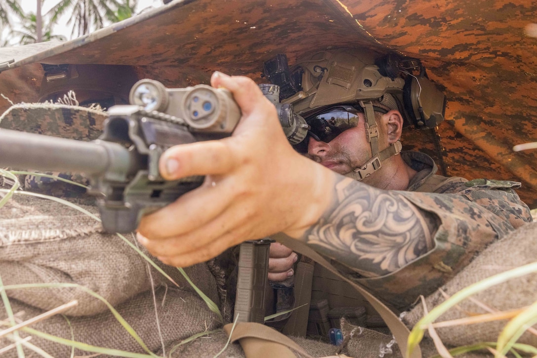 A Marine in tactical gear aims a weapon on sandbags under a camouflage tent.