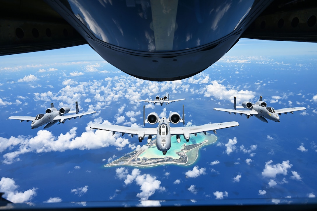 Four aircraft fly through a blue sky near clouds over an island as seen through the open exit of an aircraft.
