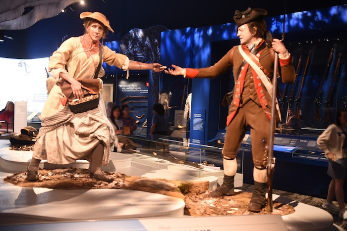 A female mannequin dressed in Colonial attire is shown passing ammunition to a male mannequin in a Continental Army uniform while on display at a museum.