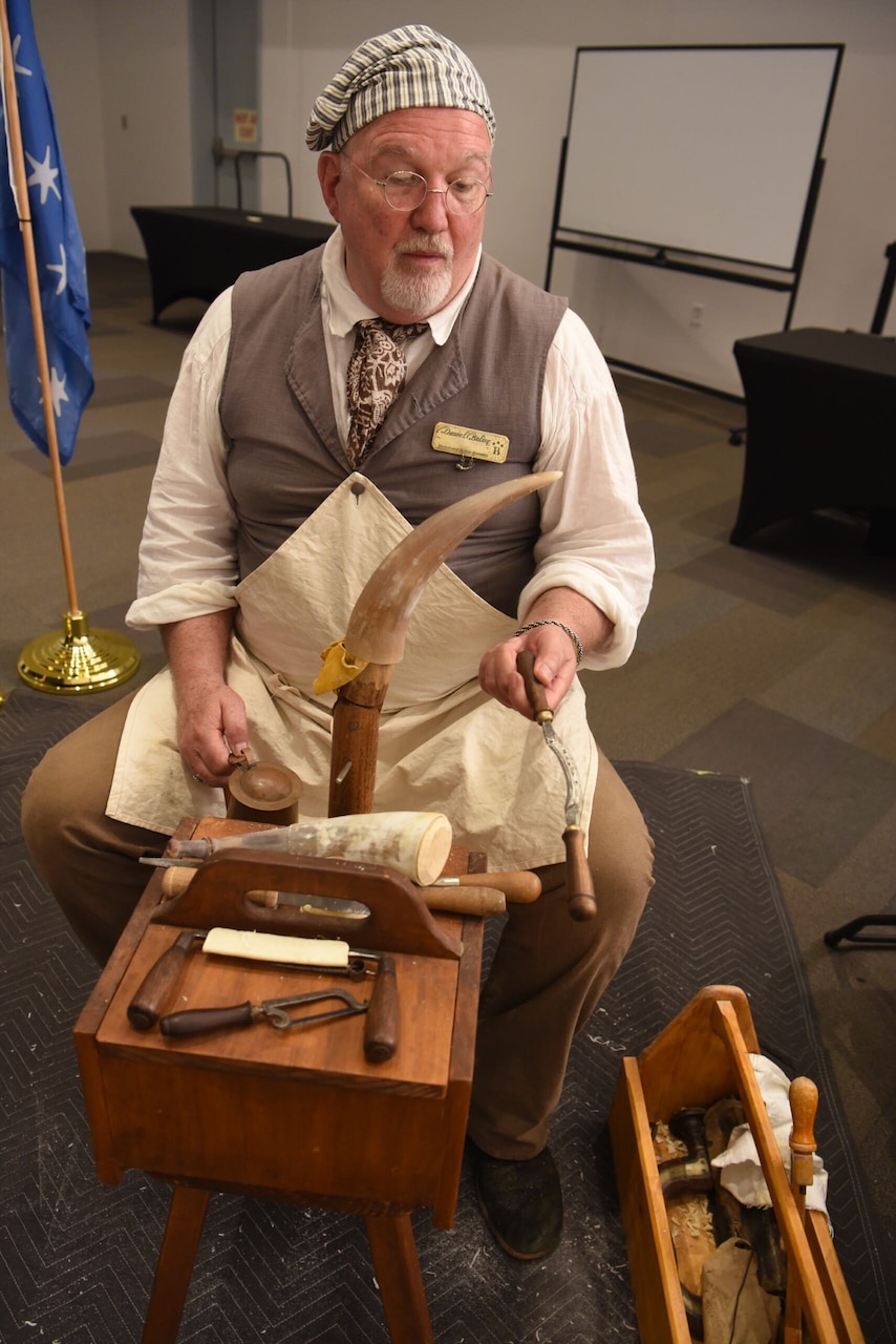 A man dressed in Revolutionary War-era clothing sits at a workbench with tools and a horn in front of him.