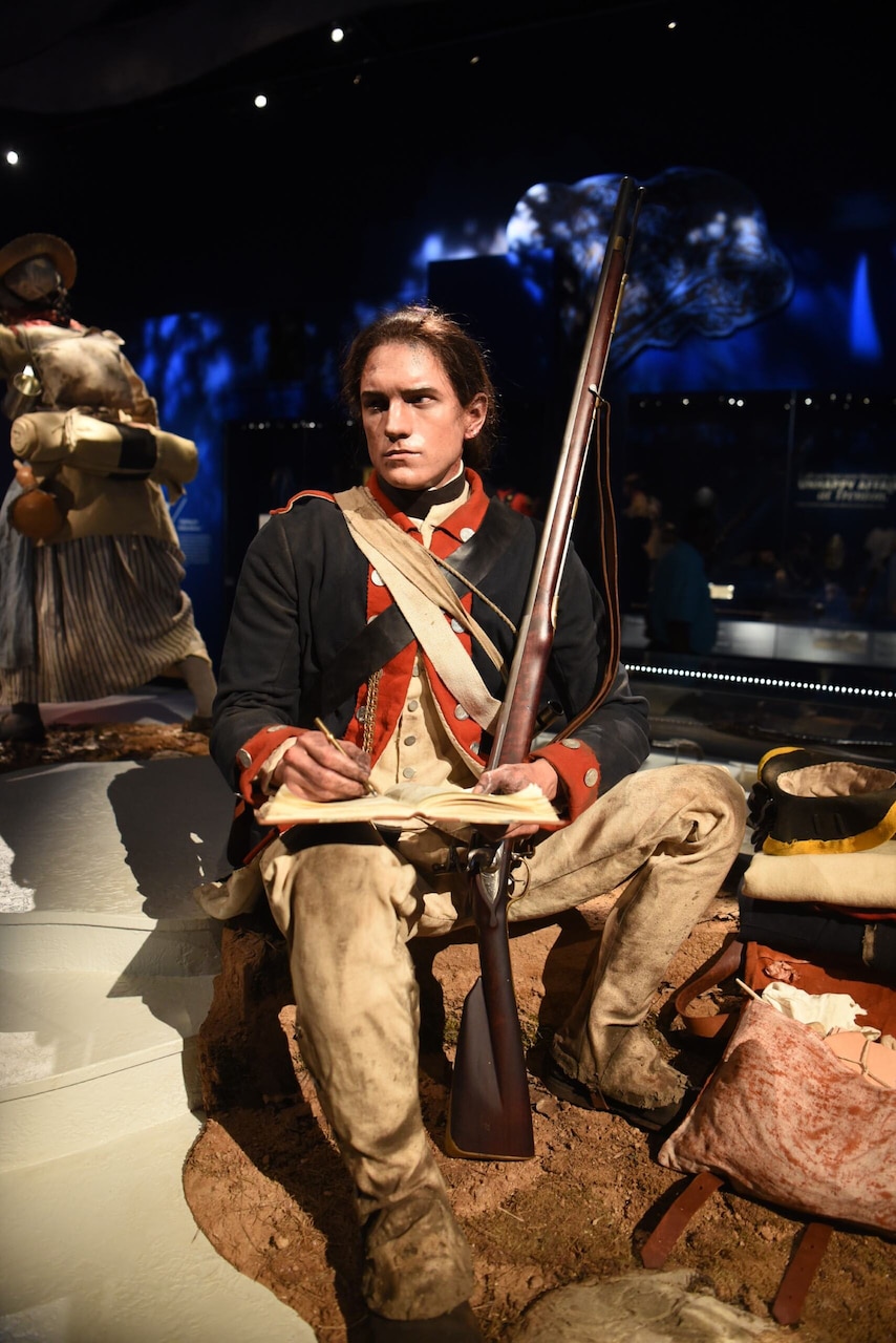 A mannequin in a Continental Army uniform is shown writing in a book with a rifle leaning against his body while on display at a museum.