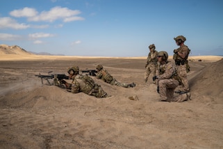 U.S. and Chilean service member stand, kneel, while others lay on their stomach holding machine guns in a desert