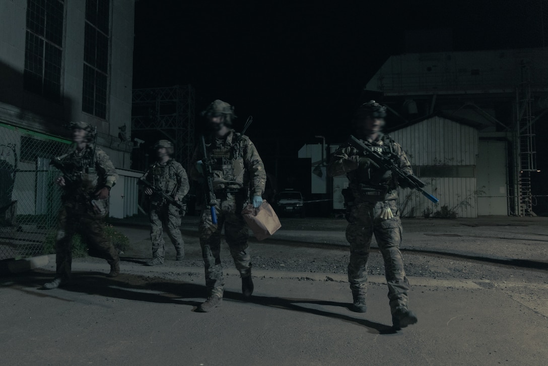 Service members carrying weapons and a paper bag walk out of a powerplant after a training