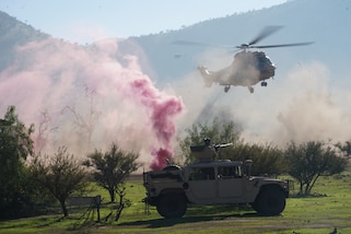 An armored military vehicle is parked in the middle of a field with a helicopter flying above it and pink smoke in the air