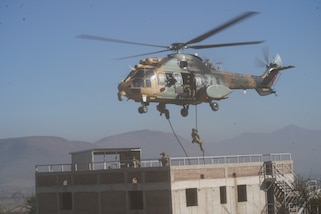 A helicopter hovers over a building while service members slide down a rope from the helicopter down to the roof of the building