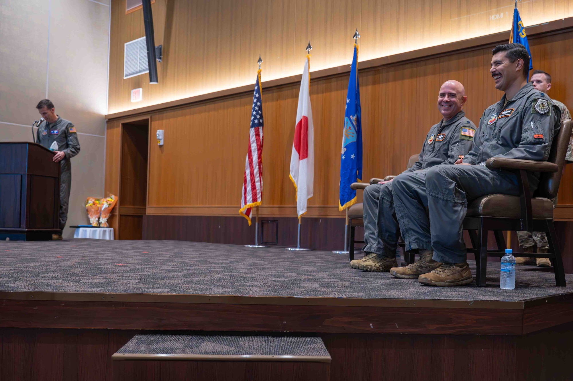 Three service members are in the photo. Two are are sitting while one is up on the podium reading his remarks.