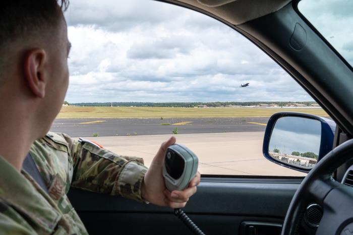 A service member holds a radio and looks out window at fighter jet taking off