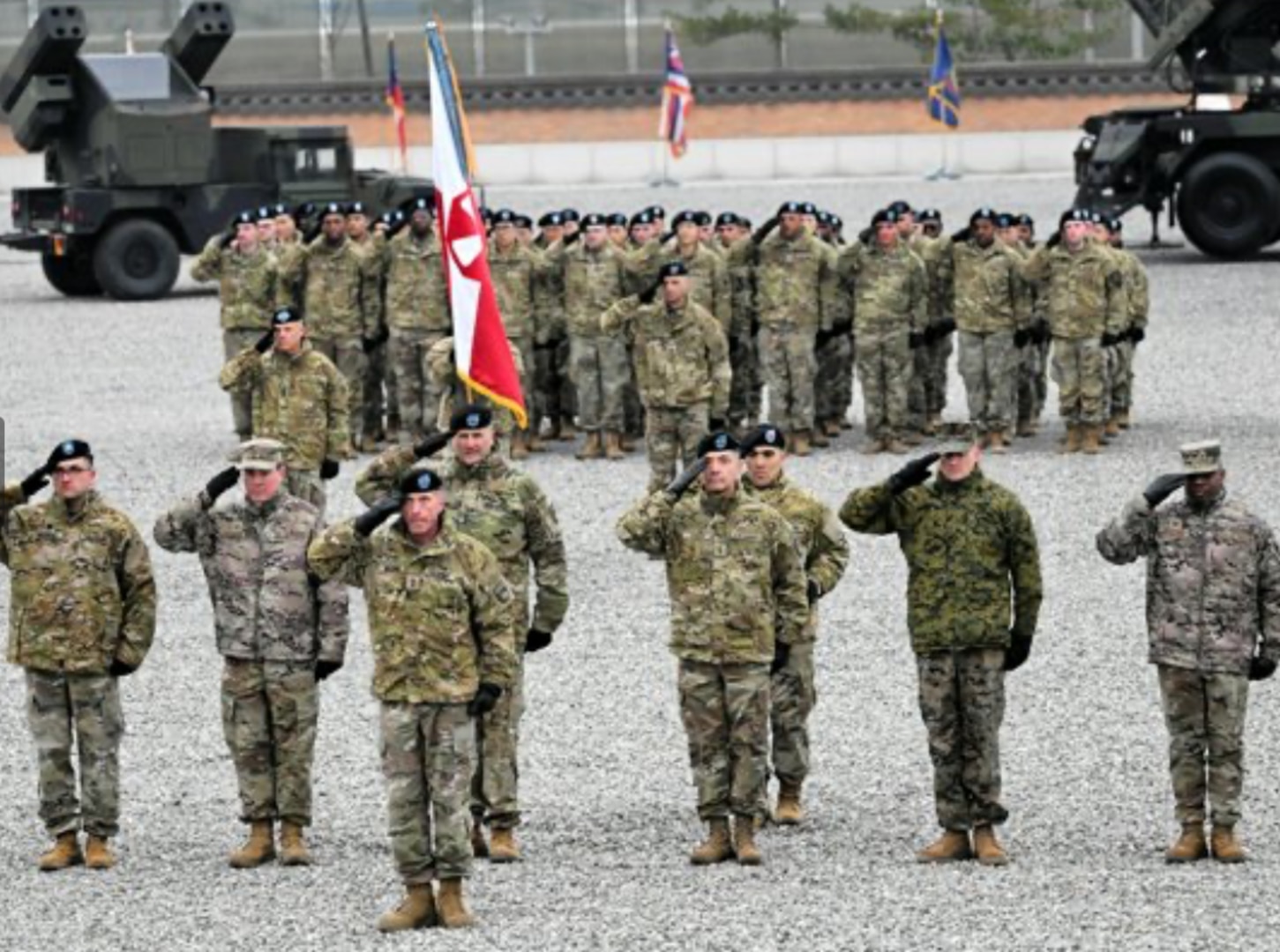 A formation of U.S. military personnel stands in uniform on a gravel parade ground, saluting during a formal ceremony. The front row consists of officers and enlisted members in various camouflage patterns, with one soldier holding a flag. Behind them, more soldiers stand in formation. Military vehicles and international flags are visible in the background.