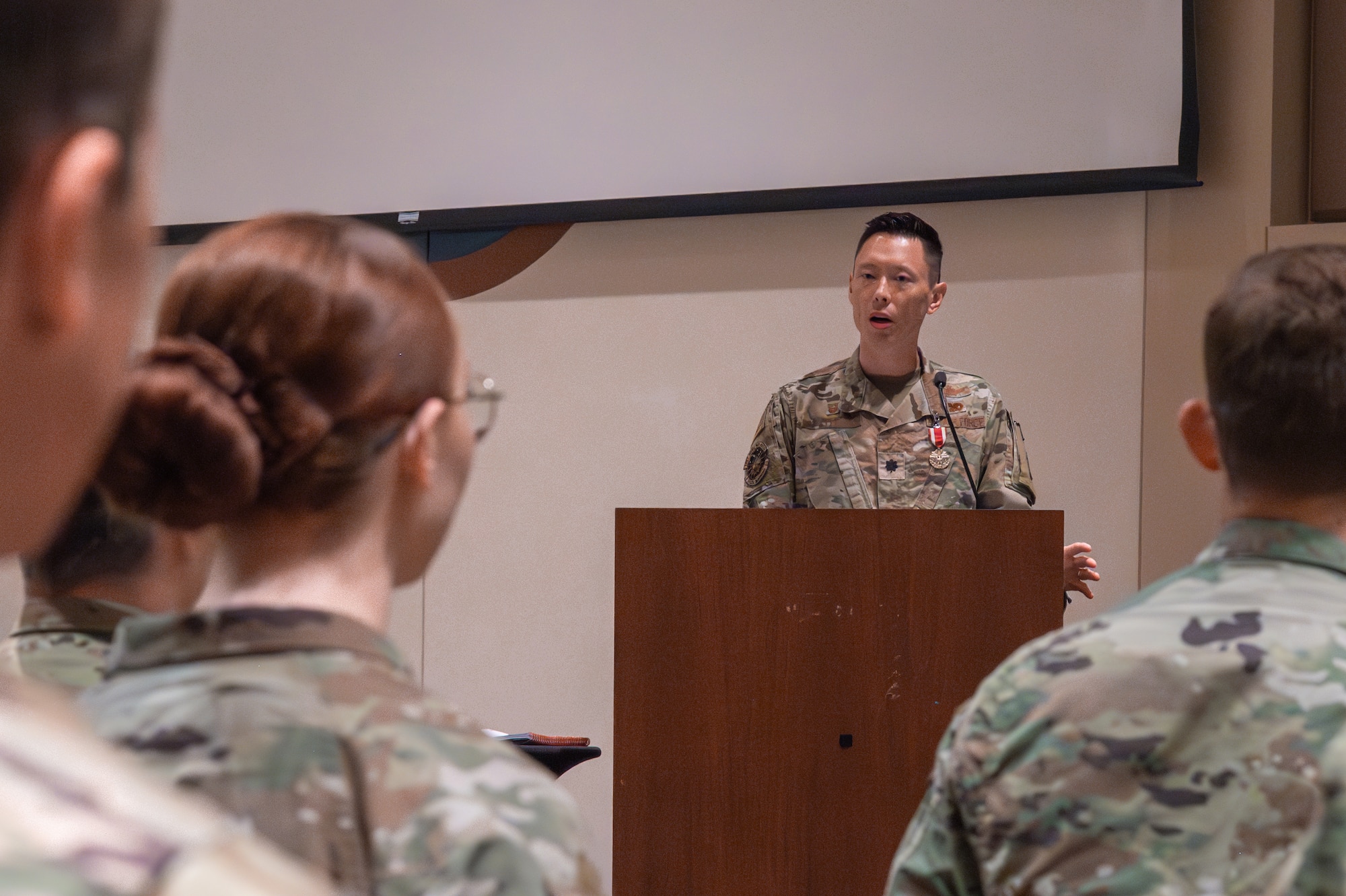 A man in a military uniform stands behind a podium speaking to a crowd of people in military uniforms.