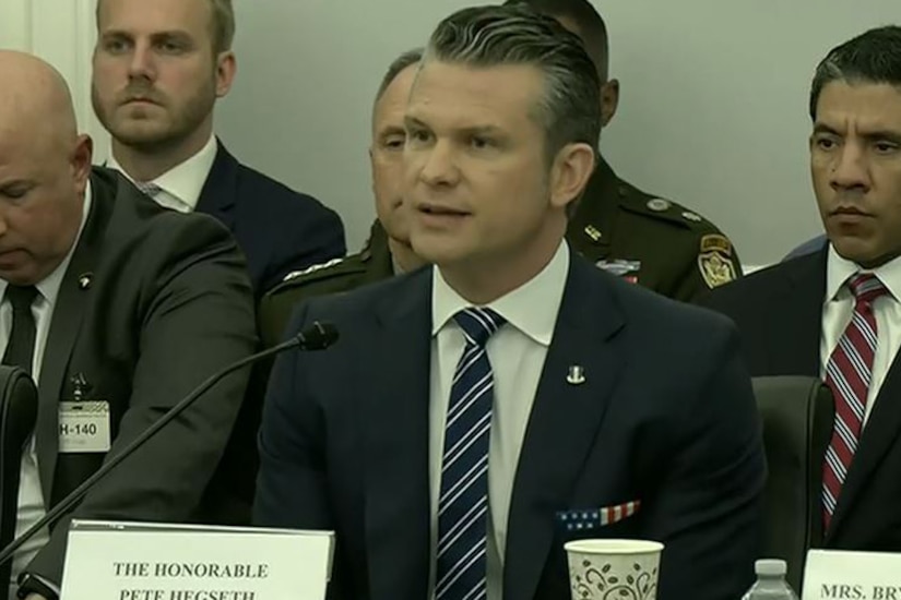 A man in a business suit is sitting at a table speaking into a microphone.