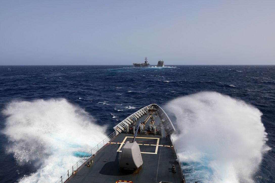 Water splashes as a partially visible ship sails toward two ships in the distance under a blue sky.