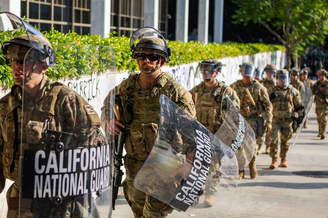 Soldiers carrying rifles and shields walk along a sidewalk on a sunny day.
