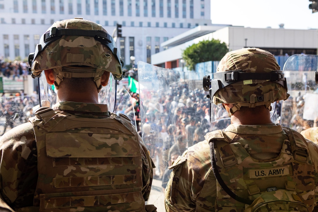 Two soldiers wearing helmets with face shields stand and watch a crowd of people gathered outdoors on a sunny day.