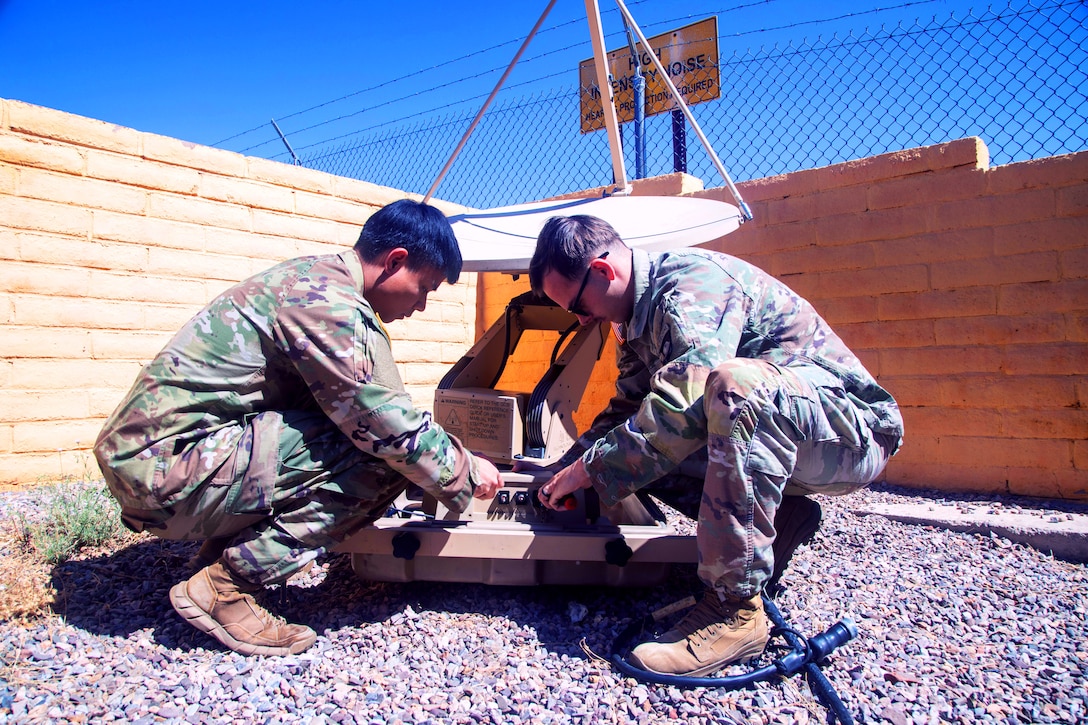 Two kneeling soldiers work on digital equipment outdoors on a sunny day with a wall, a fence and a sign in the background.