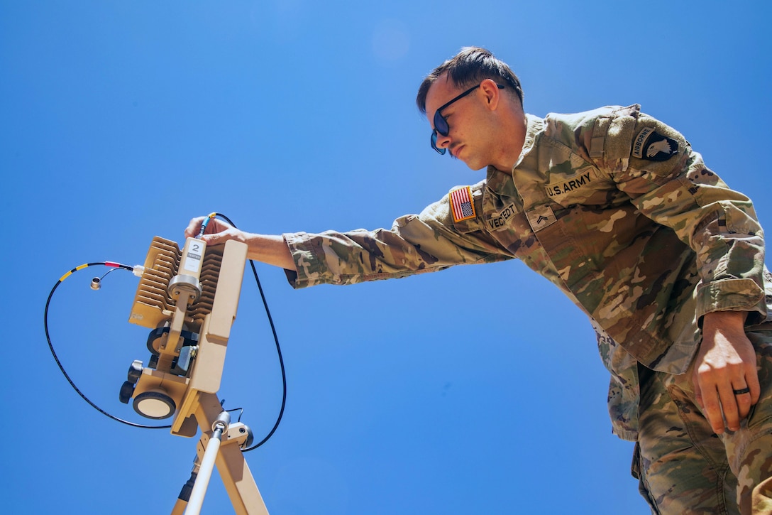 A standing soldier wearing sunglasses attaches a cable to an antenna-like device on a cloudless day.