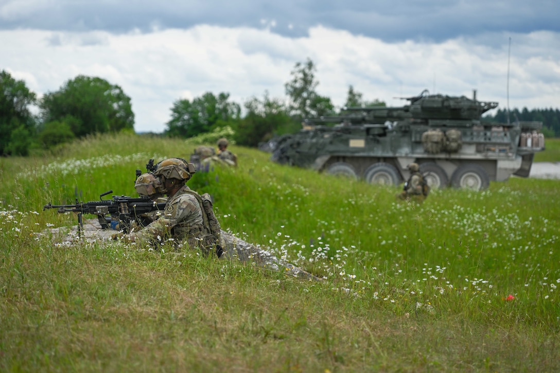 Soldiers lay on a grassy hill while aiming weapons with tanks parked to the right under a cloudy sky.
