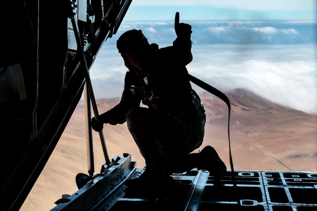 An airman kneels while holding up a finger on the ramp of an airborne aircraft with a desert and clouds in the background.