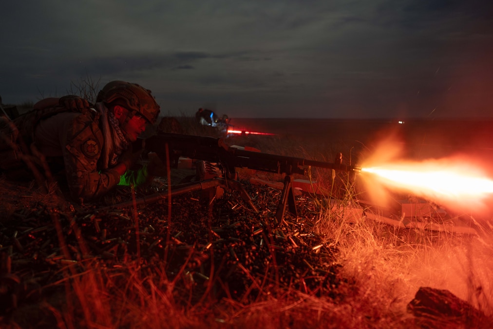 An airman lays in the grass while firing a weapon creating and orange blast under dark skies.