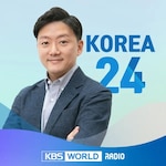 A promotional image for the radio show "Korea 24" on KBS World Radio. It features a man in a blue blazer and light shirt, smiling with arms crossed. The background has a gradient of light blue and white with wave designs. The text "KOREA 24" is prominently displayed next to him, and the KBS World Radio logo is at the bottom.