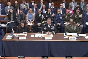 A woman wearing business attire sits at a desk and speaks into a microphone; two men wearing military uniforms are seated next to her.
