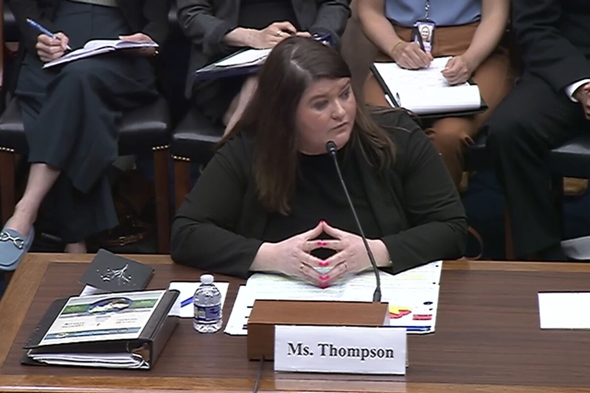 A woman wearing business attire sits at a desk and speaks into a microphone.